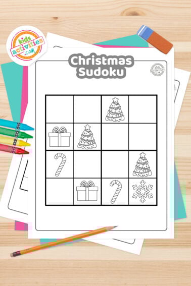 Black and white Christmas sudoku printable with Halloween-themed icons on top such as christmas tree, a gift, a snowflake and a candy cane; on top of blue-green and purple sheets with assorted crayons on a light brown background.