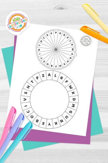 Black and white cipher wheel printables for kids to write their own codes, on top of blue-green and purple sheets with assorted crayons on a dark grey background. printed pdf version from Kids activities blog.