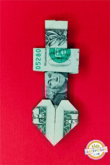 Finished result of a dollar bill origami heart with cross done on top of a red background. tutorial from kids activities blog