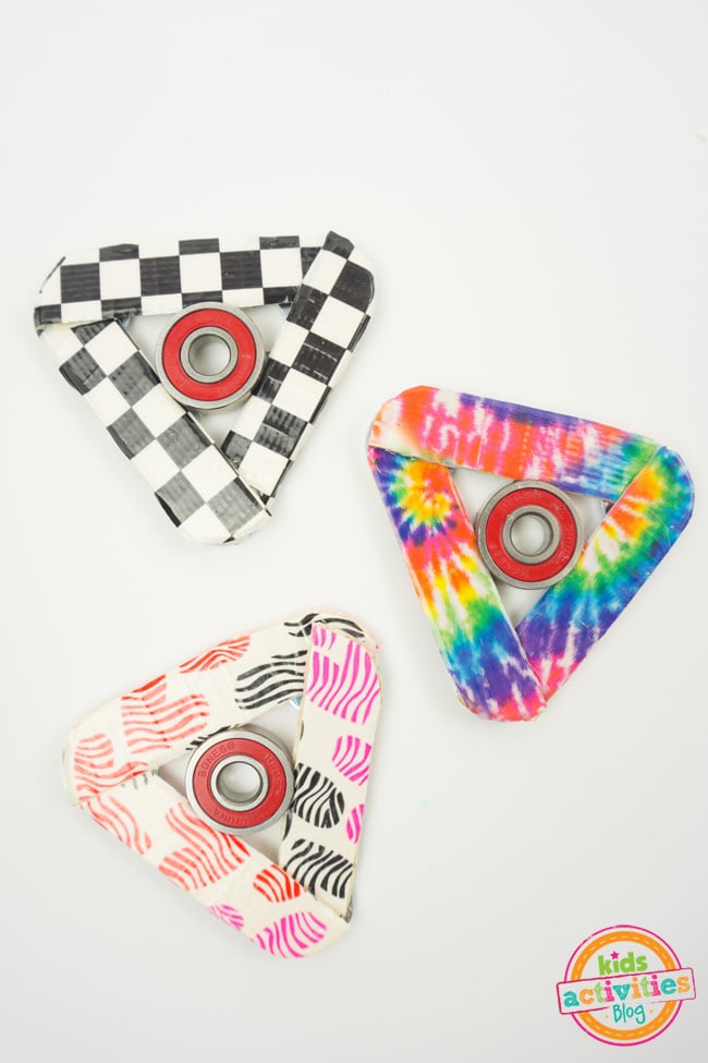 How to Make Fidget Spinner craft - three completed homemade fidget spinners shown - one with checker board tape, one decorated with rainbow tie dye tape and one with multi-color hearts