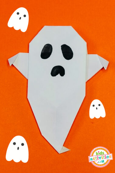 White origami ghost done with a drawn-in face on top of an orange background, and ghost doodles for decoration.