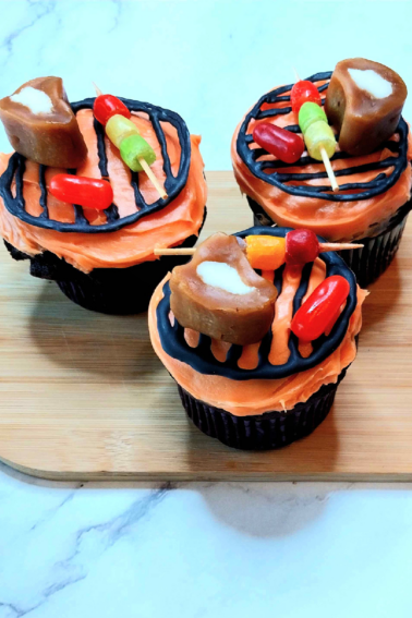 BBQ Cupcake- 3 Finished Cupcakes with orange icing, black grill candy, and candies that look like meat and veggies on a cutting board- Kids Activities Blog