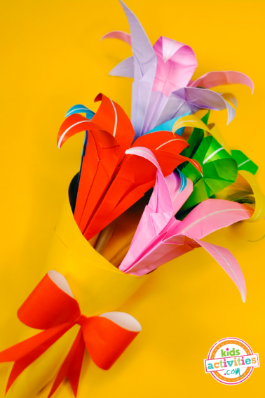Image shows an origami flower bouquet made with flowers in different colors, tied with a red ribbon, on top of a yellow background
