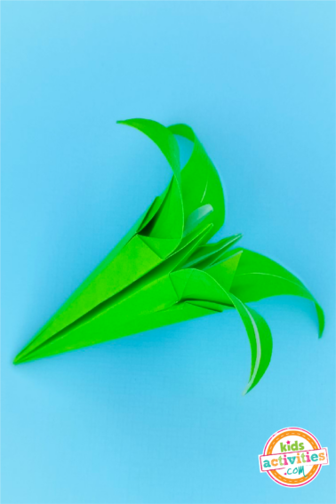 Final step of an origami lily made with green origami paper on top of a blue background.