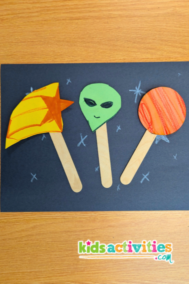 Outer space art activity. Image of black paper with white stars, and three paper puppets on popsicle sticks. One is a shooting star, one is an alien, and one is an orange planet. - kidsactivities.com